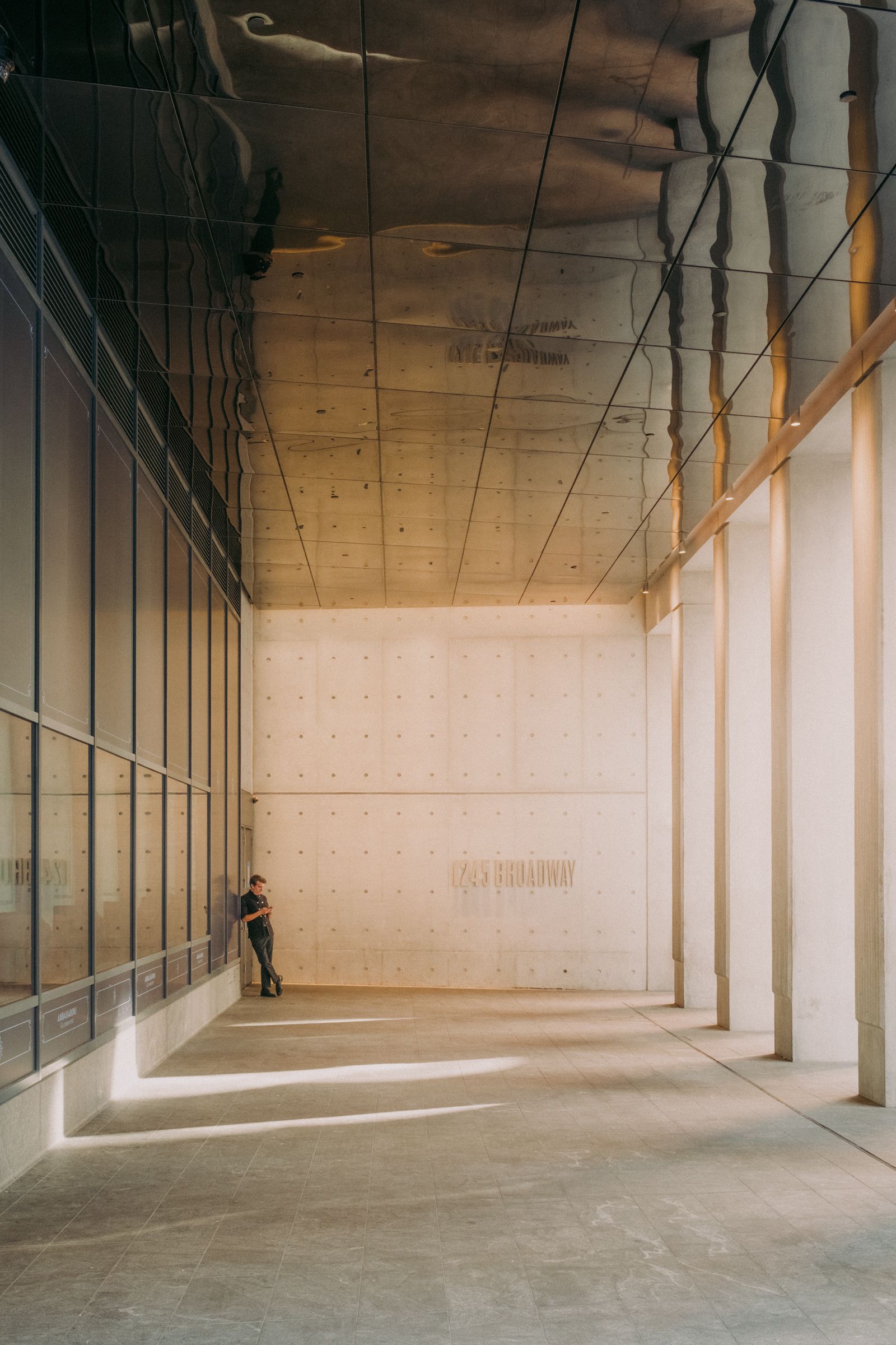 A lone figure stands against the far wall of a tall, empty concrete colonnade, dwarfed by the columns; the building’s address is embossed on the back wall and a mirrored soffit overhead reflects the street outside.