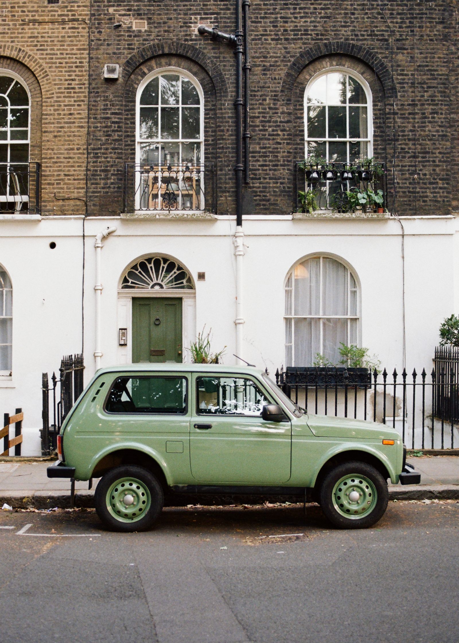 A pale green vintage 4x4 parked on a city street in front of a brick and white stucco townhouse with arched windows.