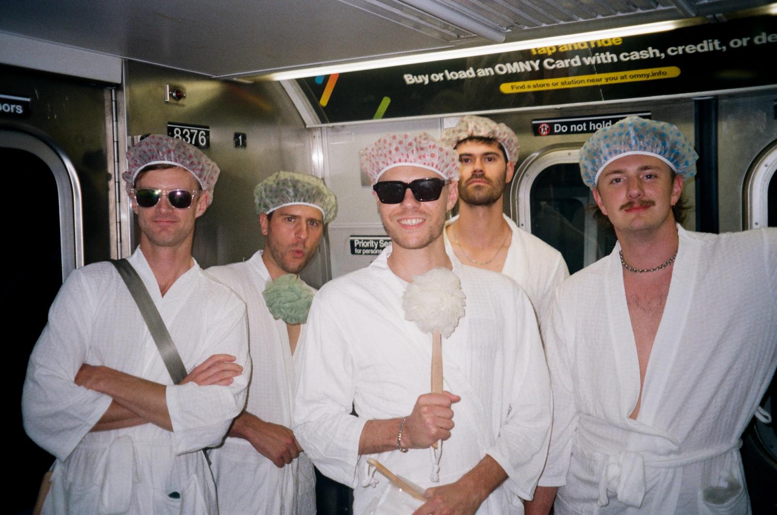 Five men in white bathrobes, shower caps and sunglasses crowd together grinning on a subway car, one holding a loofah on a long handle.