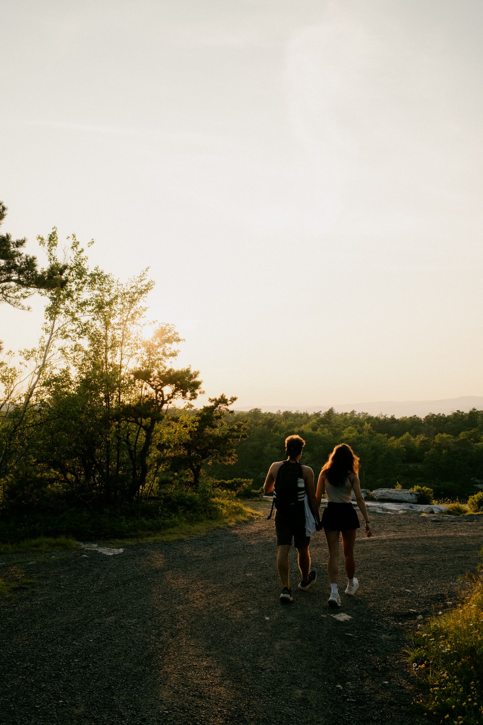 A couple stands on a rock outcrop above a forested lake at sunset, turned toward each other and smiling, a daypack slung over his shoulder.