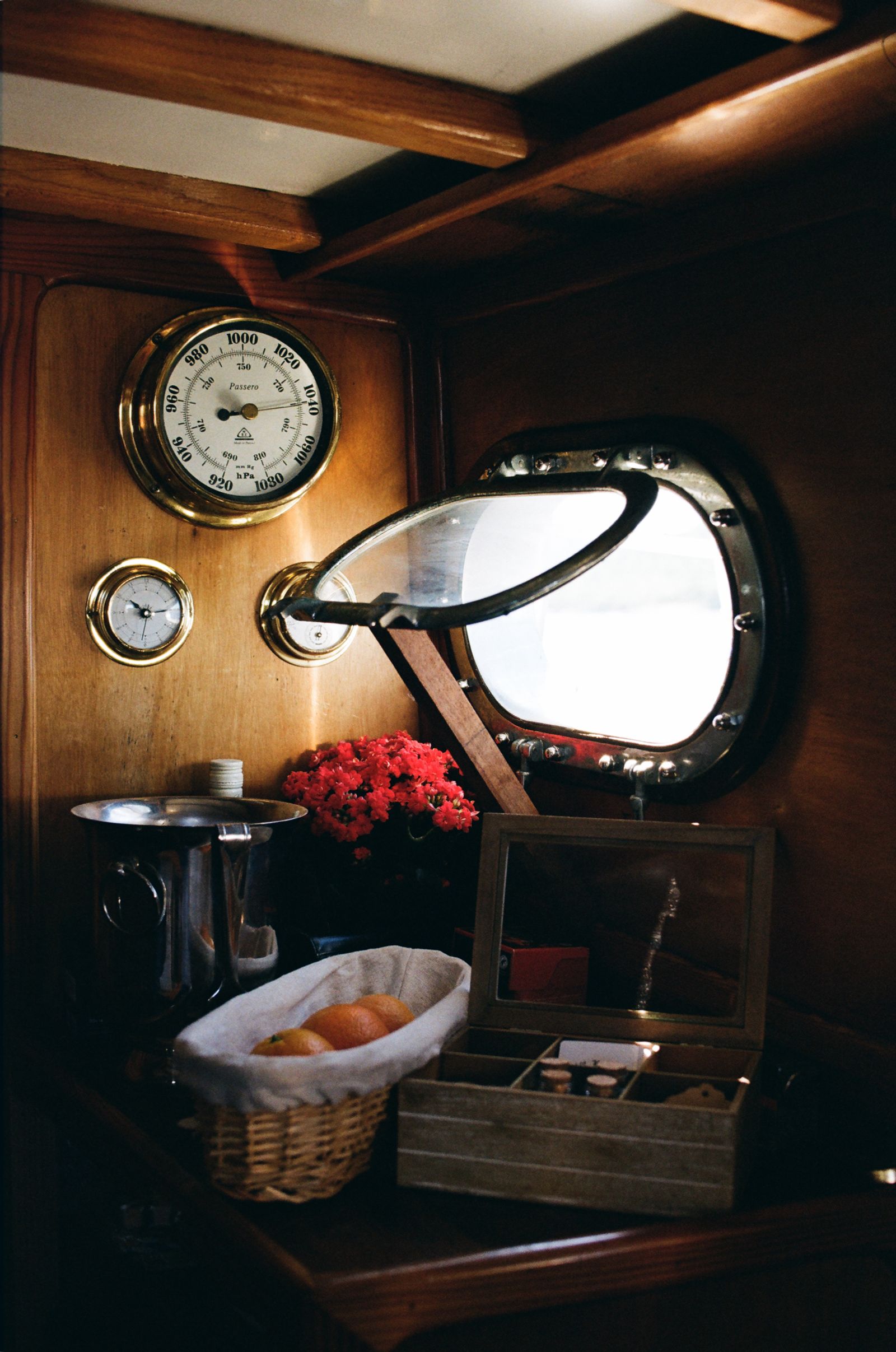 Brass barometer and porthole in a wood-paneled boat cabin, with red flowers, oranges, and a small trinket box below.