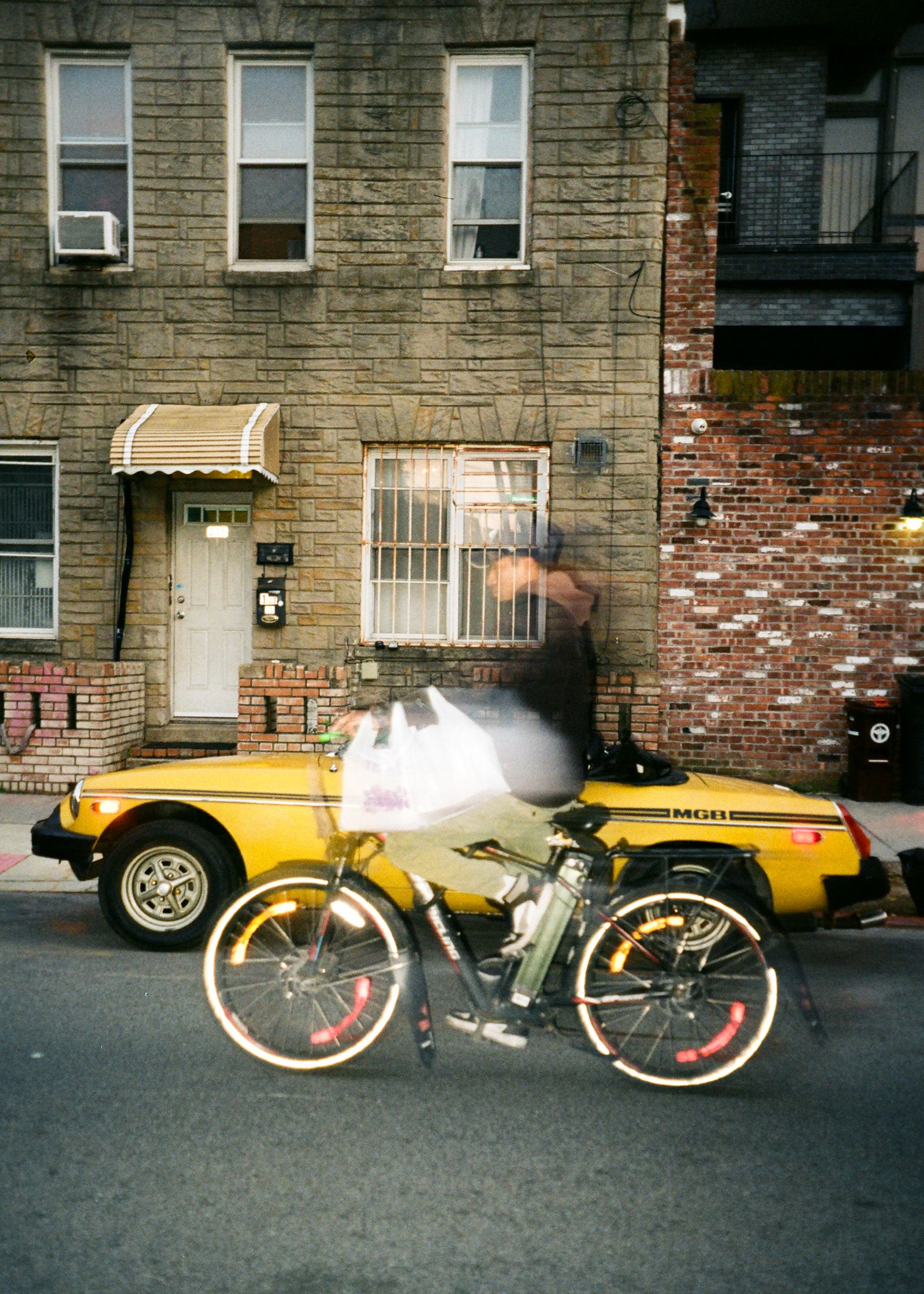 A motion-blurred cyclist passes a parked yellow vintage MGB convertible on a city street lined with brick rowhouses.