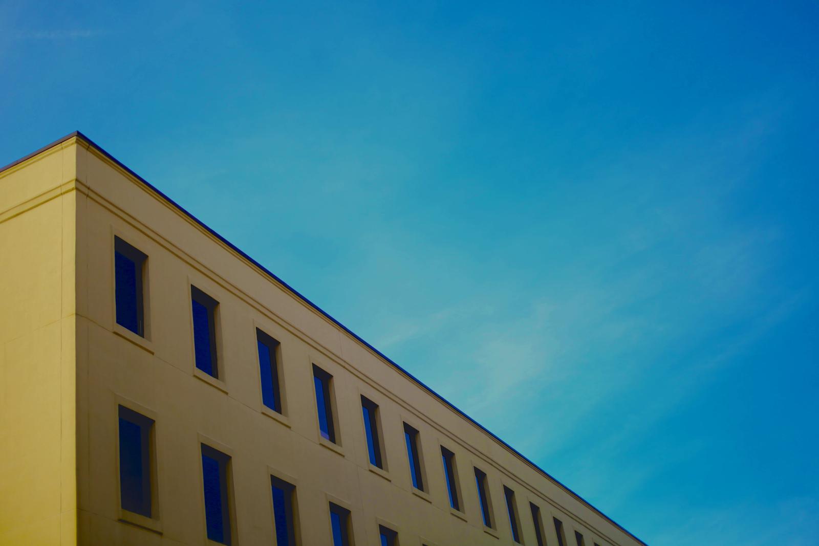 The sunlit mustard-yellow corner of a building with rows of windows angles into a clear blue sky.