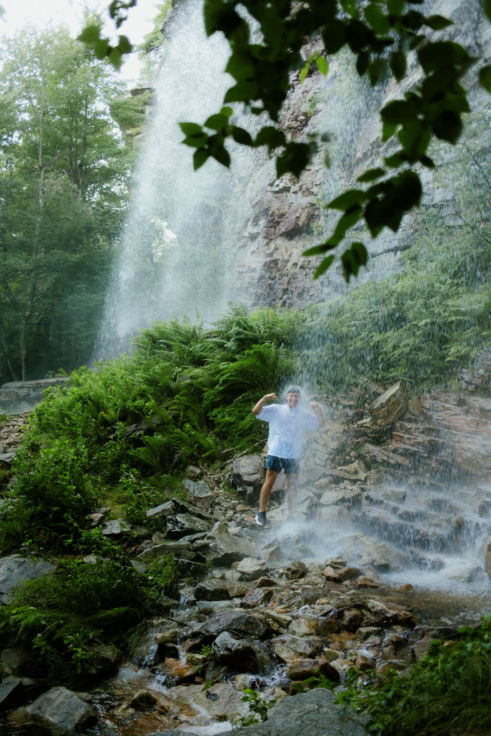 A man stands beneath a rushing waterfall in a fern-lined ravine, arms raised and flexed, grinning as spray drenches his white t-shirt and shorts.