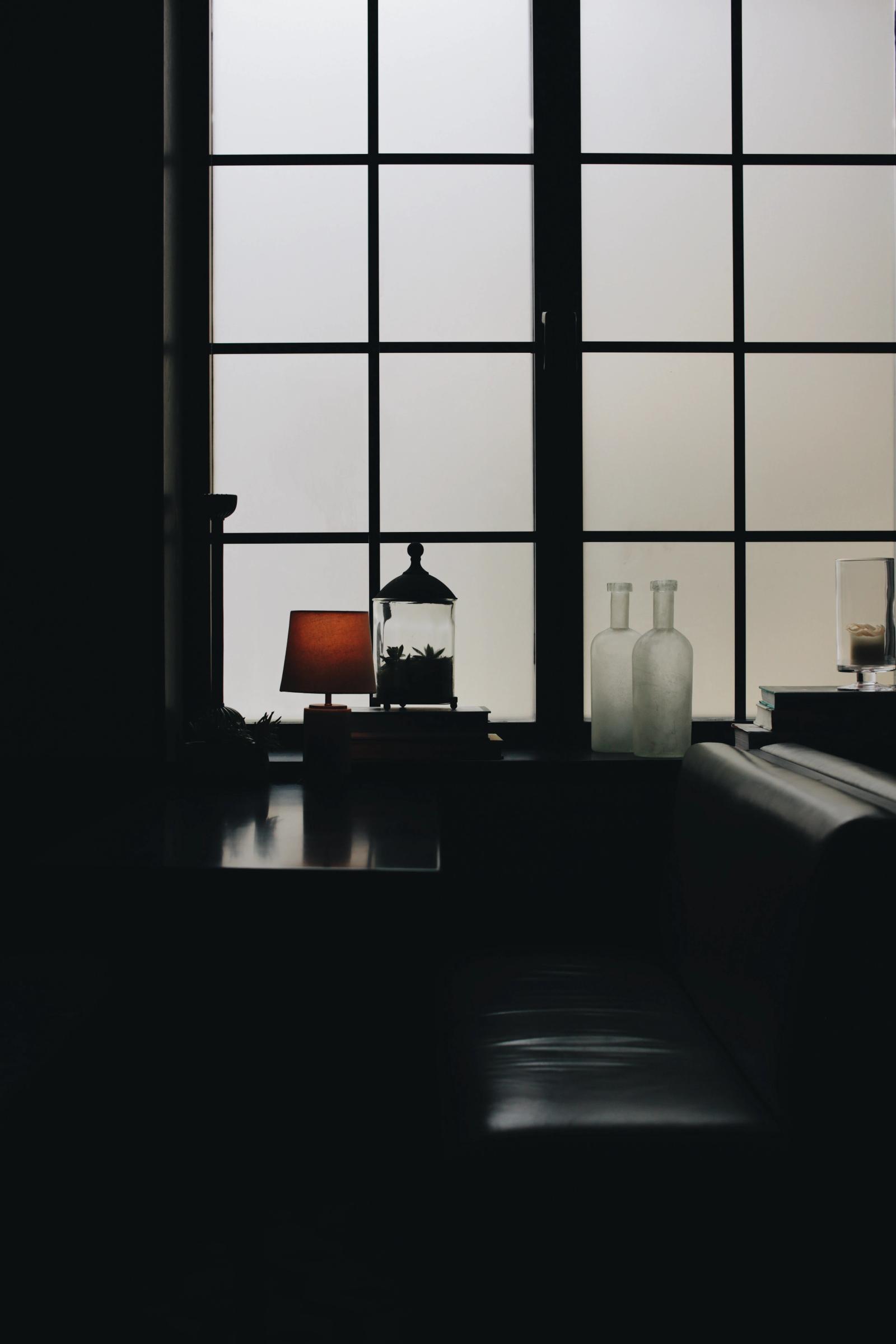Silhouetted lamp, glass jar, and bottles arranged on a windowsill against frosted window panes at dusk.