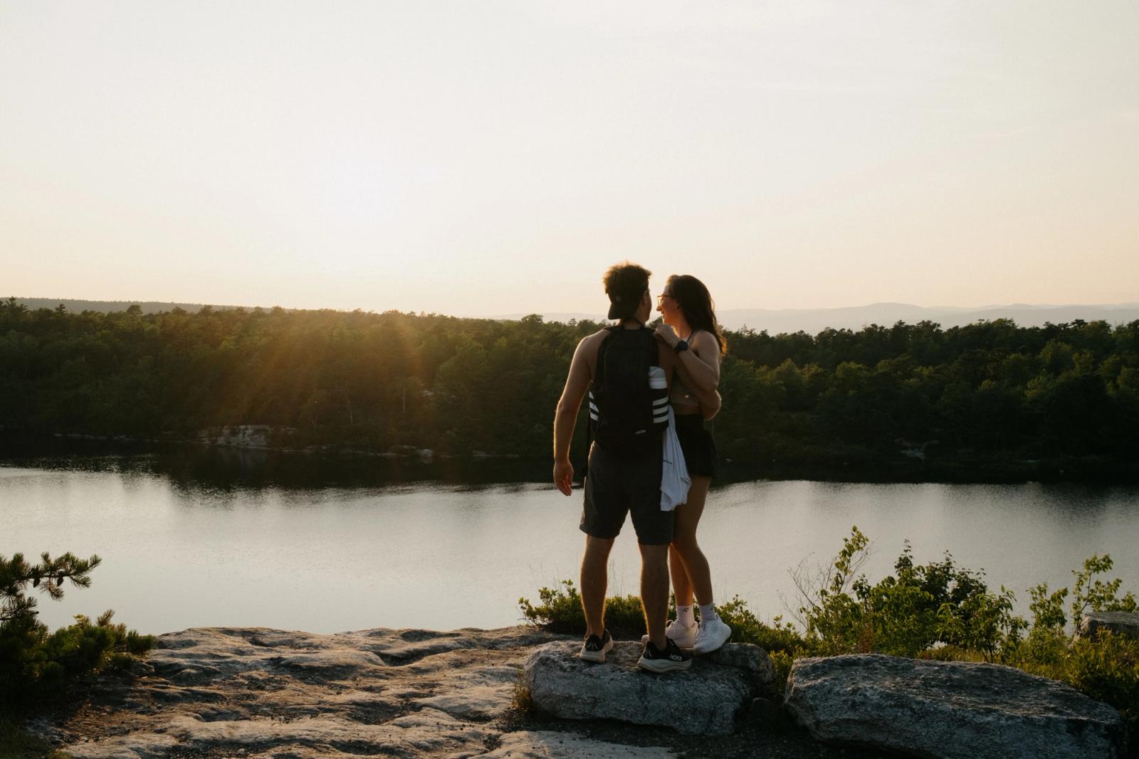 A couple walks hand in hand away from the camera along a rocky ridge trail, silhouetted against a hazy sunset over the treeline.