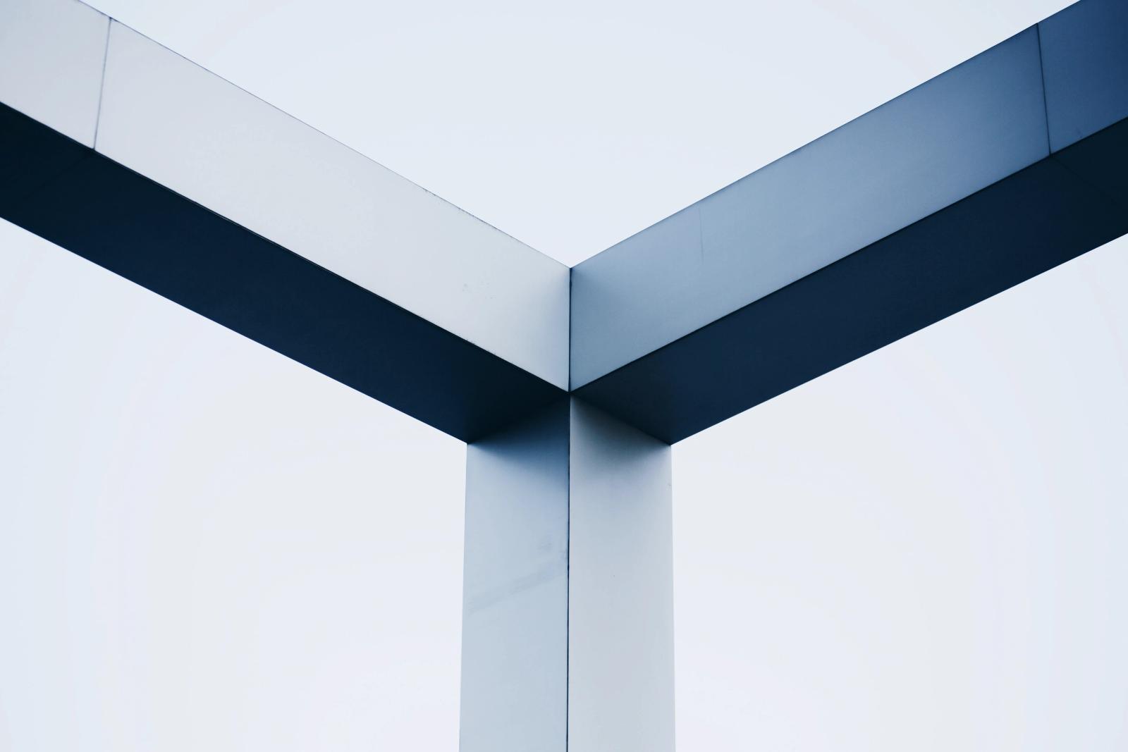 Black-and-white close-up of two angled steel beams meeting a vertical column against a pale sky.