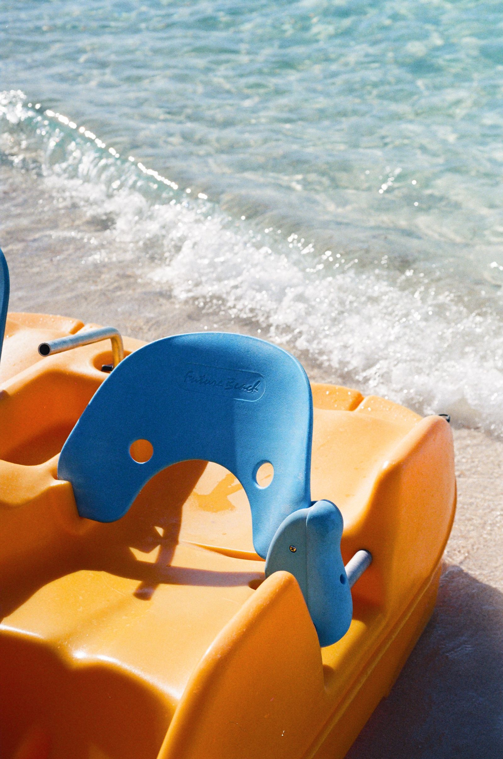 Close-up of an orange and blue pedal boat parked on sand as clear waves wash in behind it.