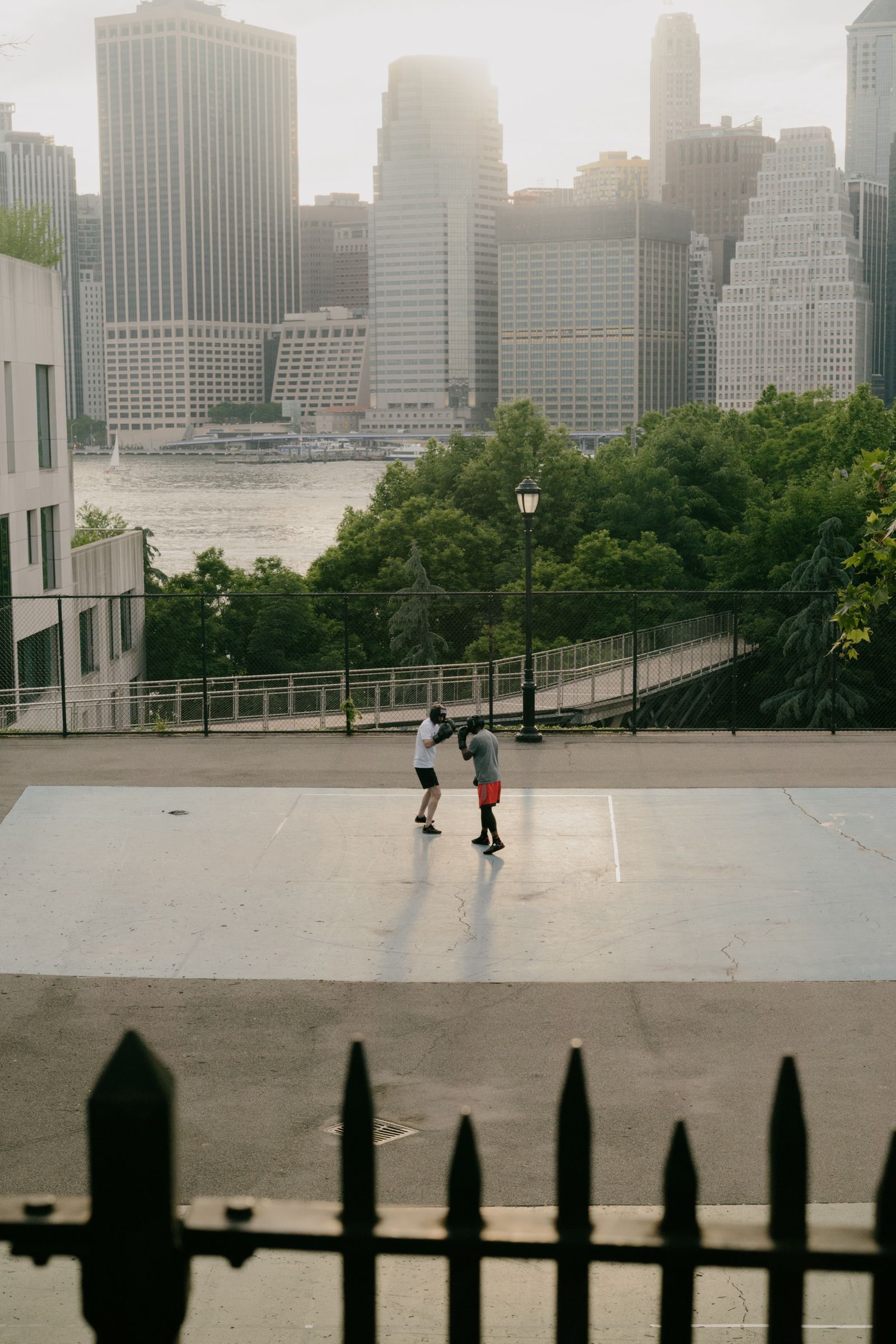 Two people in boxing gloves spar on an outdoor court with a hazy city skyline and river visible behind them.