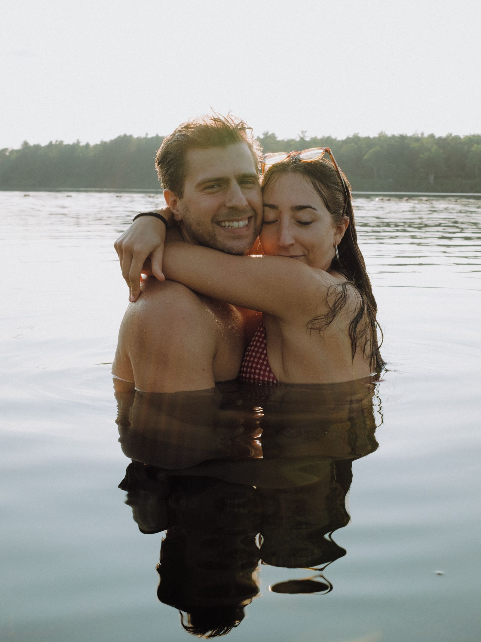 A man and woman embrace shoulder-deep in a still lake at golden hour, her sunglasses pushed back on her wet hair, both smiling with eyes closed.