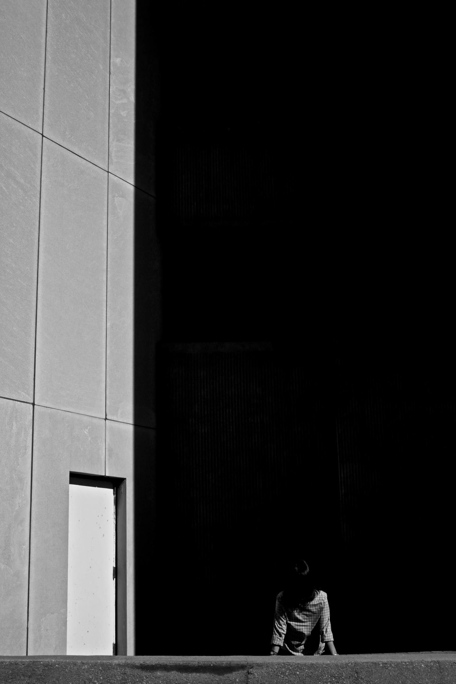 A person sits in deep shadow at the base of a concrete wall beside a brightly lit open doorway.