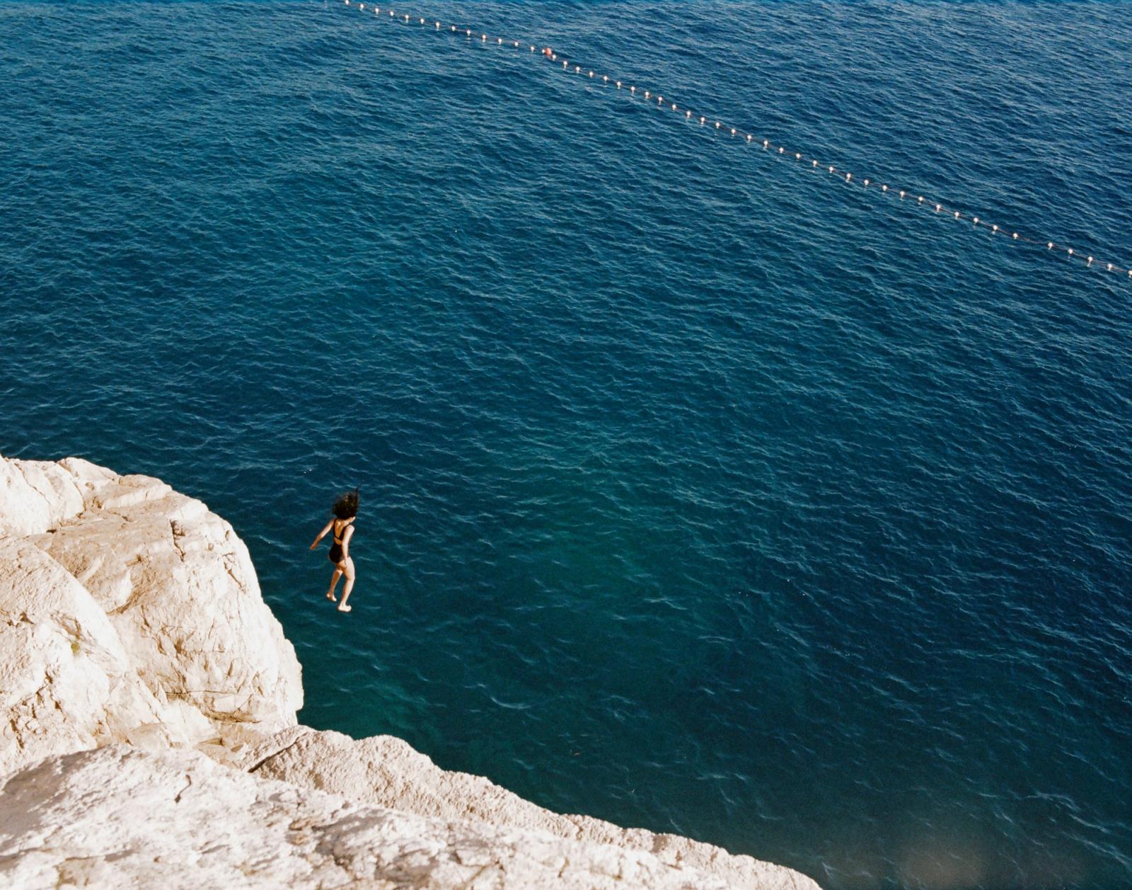 A woman in a black swimsuit leaps feet-first off a pale cliff into deep blue sea water below.