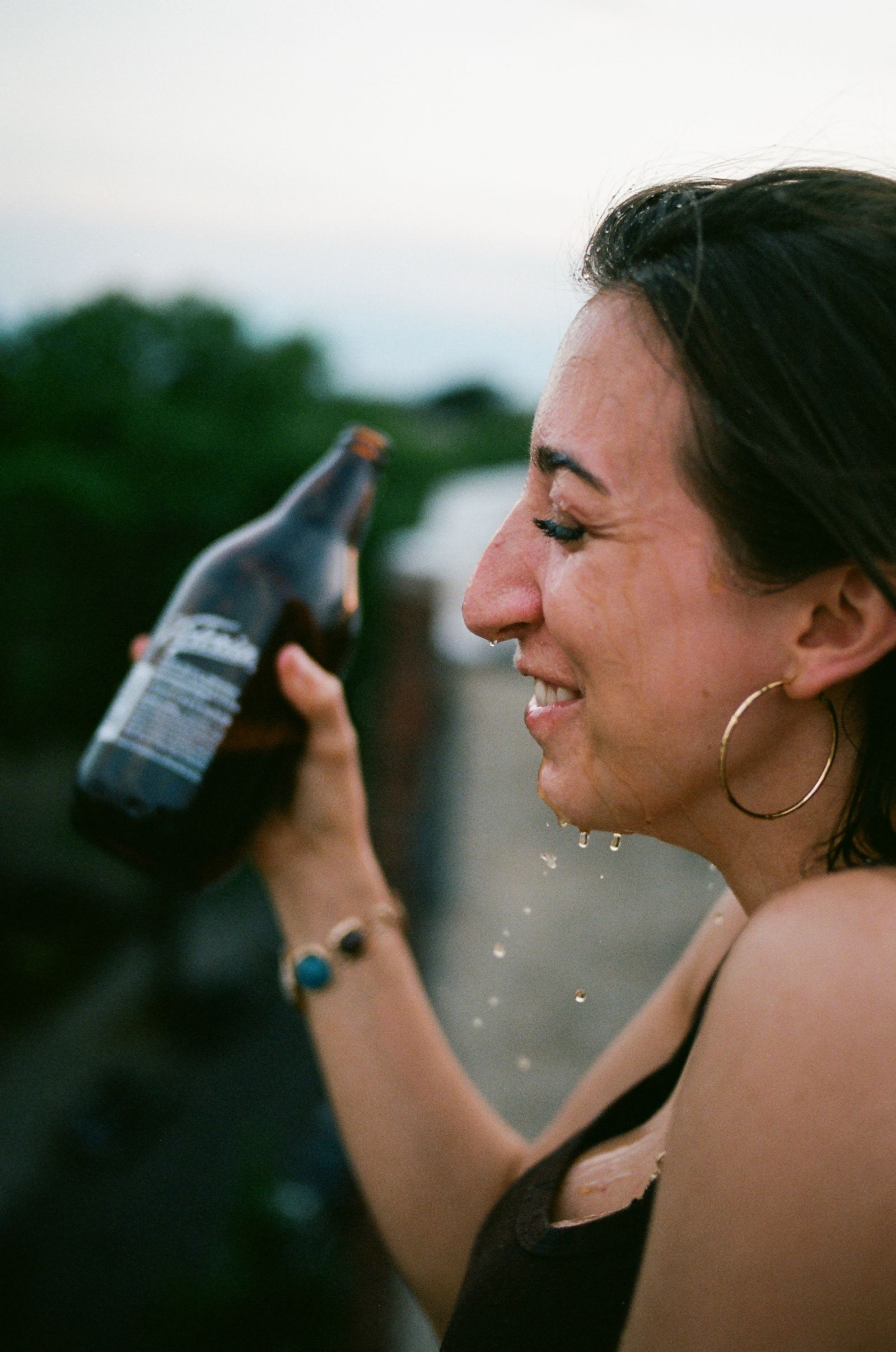 A woman laughs mid-sip from a bottle on a rooftop at dusk, droplets catching the light around her.