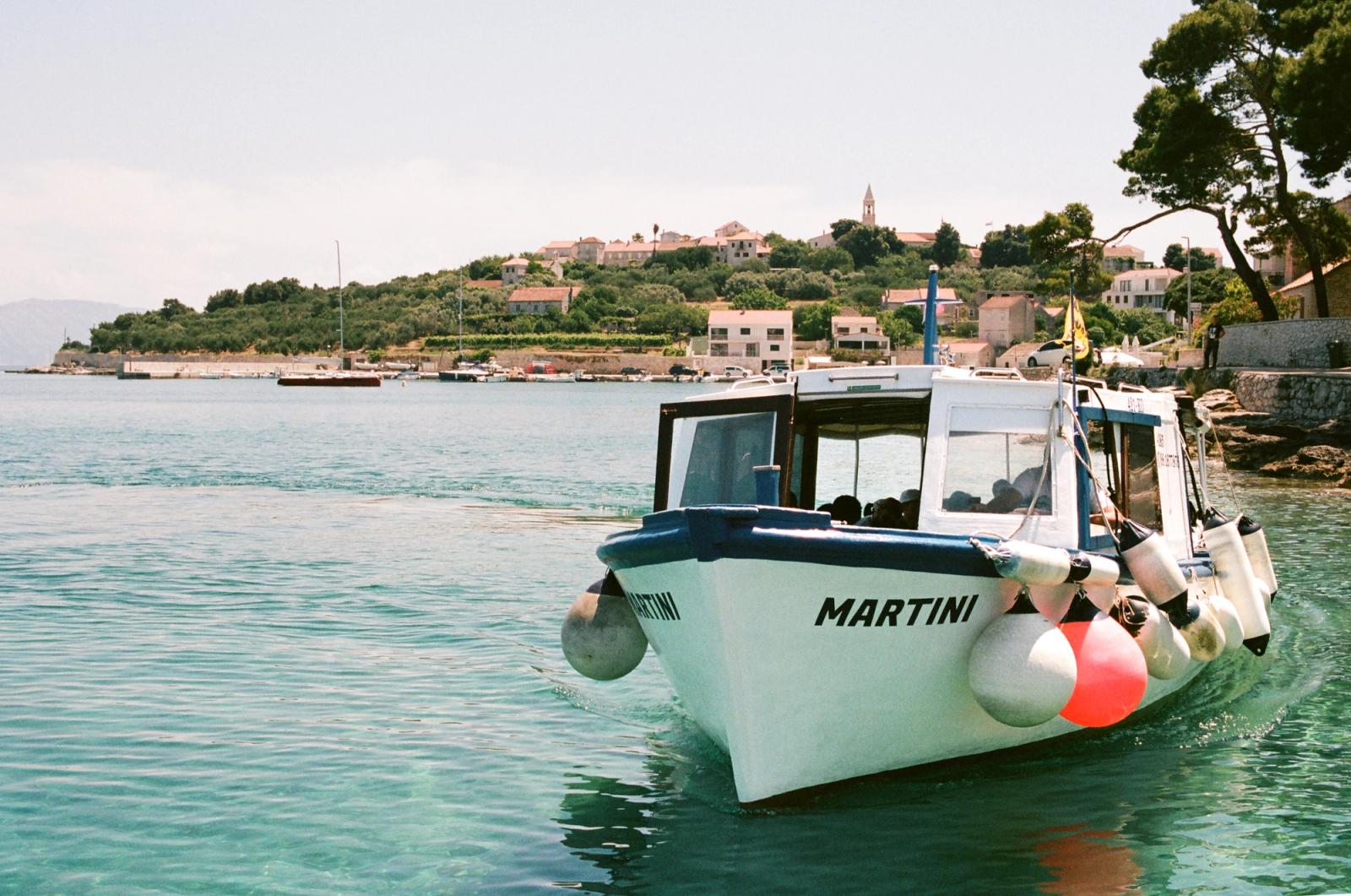 A white passenger boat named "Martini" motors toward camera past a hillside coastal village with a church bell tower.