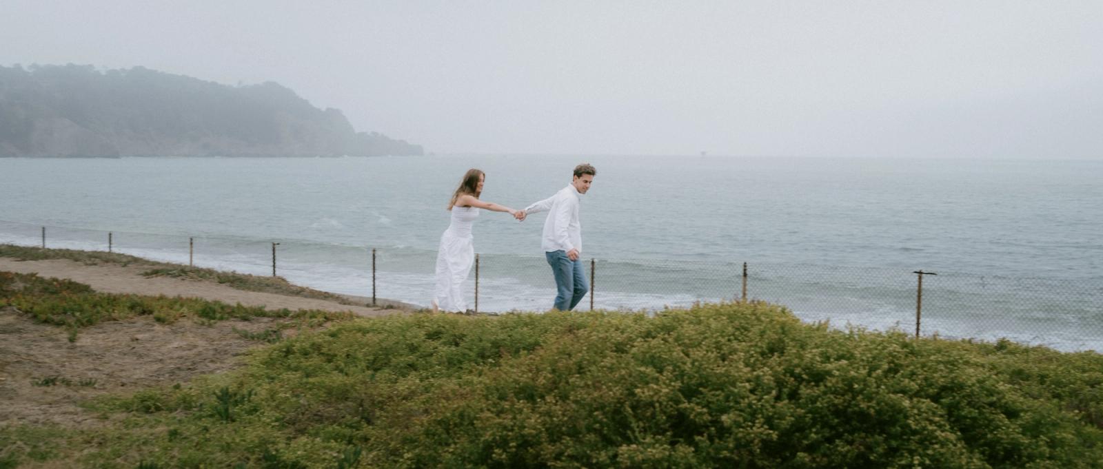 A wide panoramic frame of a couple in white walking hand in hand along a low bluff above a grey, fogbound bay.