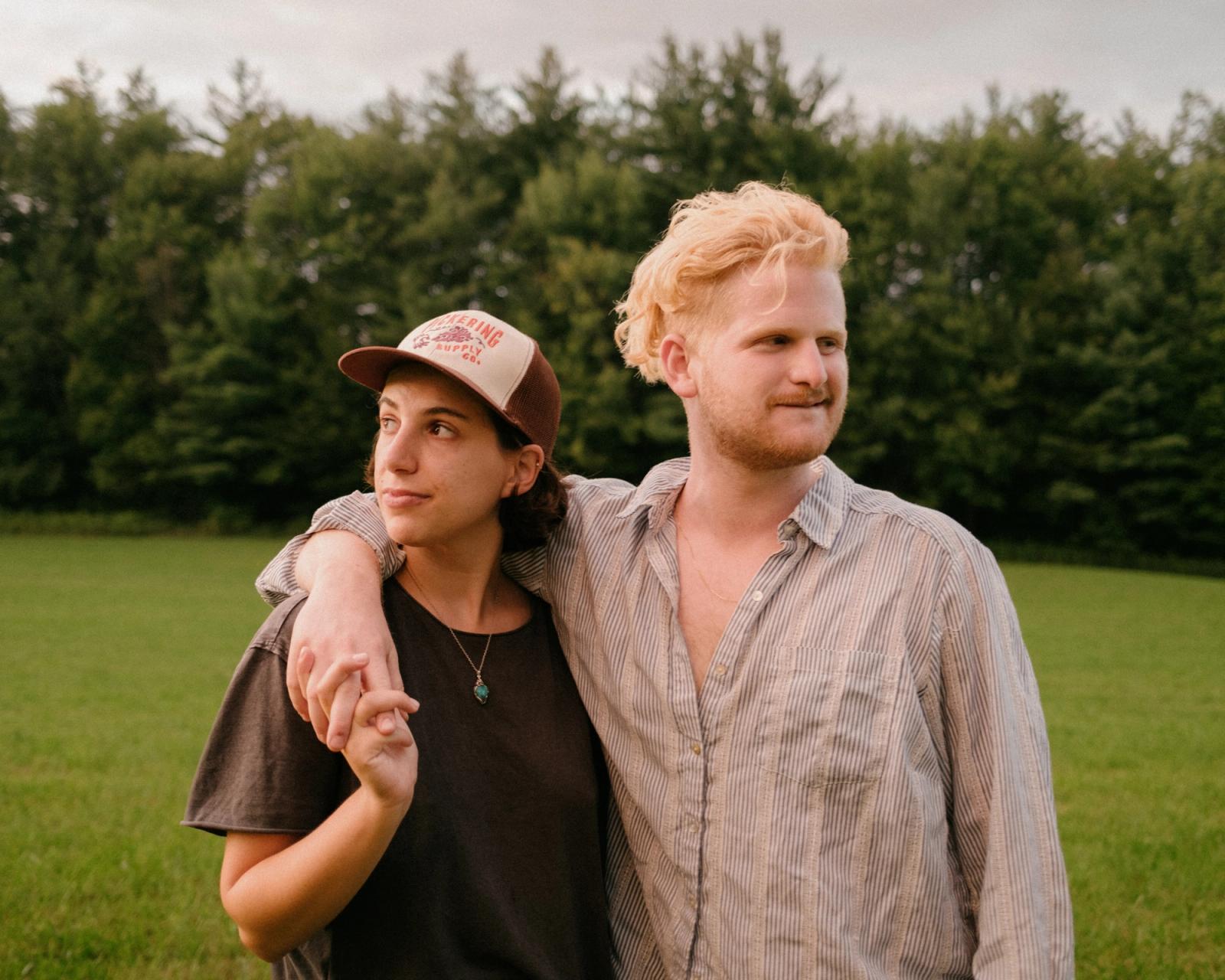A woman in a cap and a fair-haired person in a striped shirt stand arm in arm in a mown field, both looking off to one side, dark pines behind them.