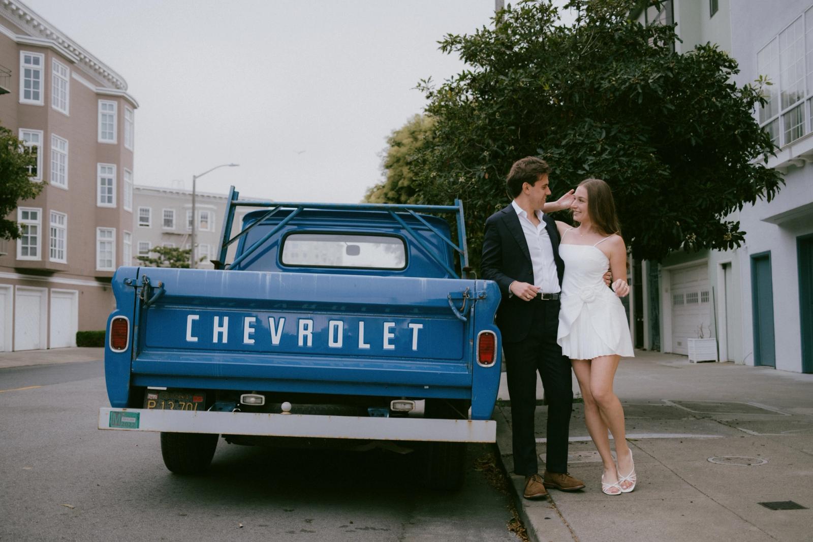 A couple in a dark suit and a short white dress lean against the tailgate of a vintage blue Chevrolet pickup on a quiet residential street, looking at each other.