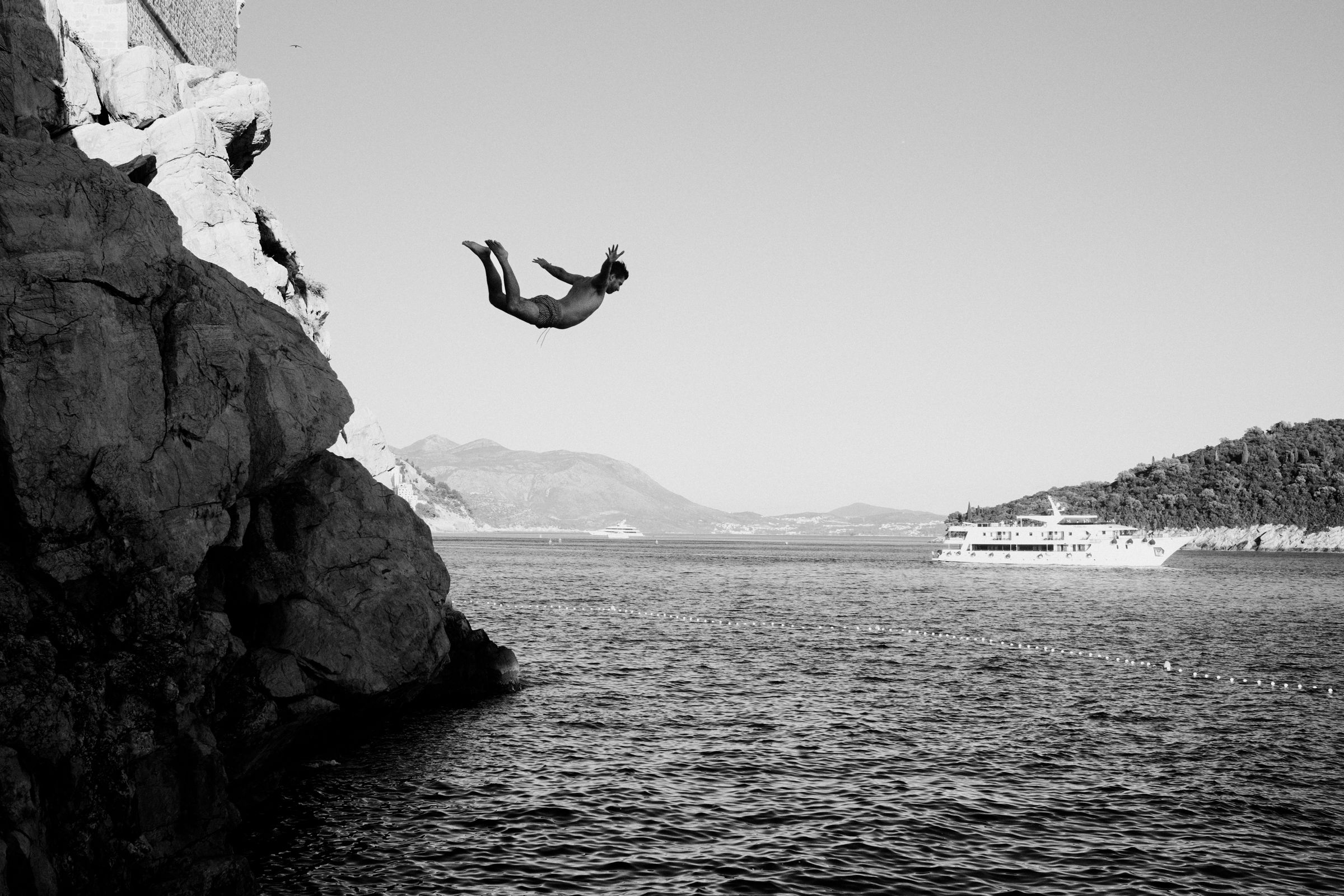 Black-and-white shot of a man mid-air backflipping off a cliff above the sea, with a passenger boat and hills behind.