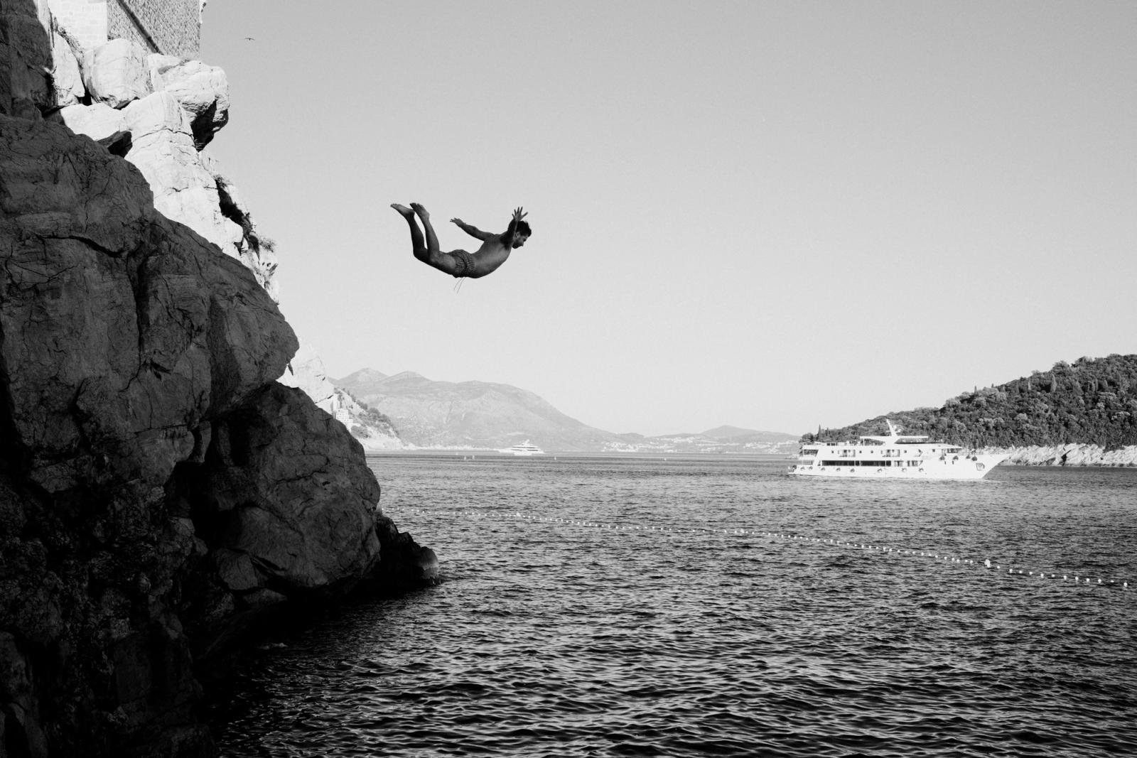 Black-and-white shot of a man mid-air backflipping off a cliff above the sea, with a passenger boat and hills behind.
