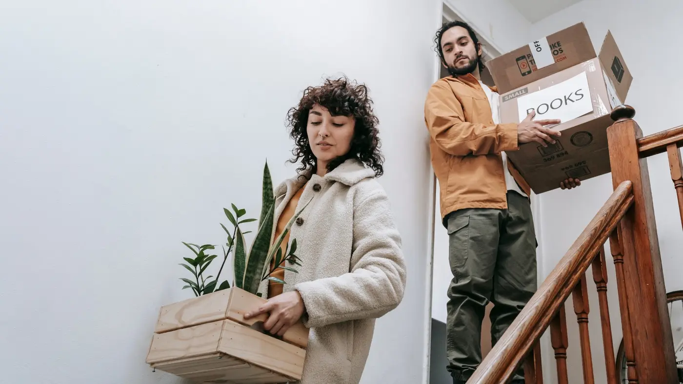 Couple carrying moving boxes and a potted plant while moving household belongings to a new home.