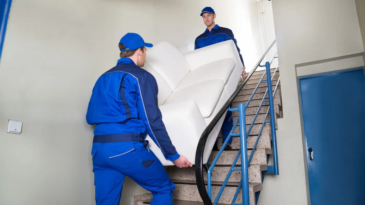 Two professional movers carrying a white sofa up a staircase.