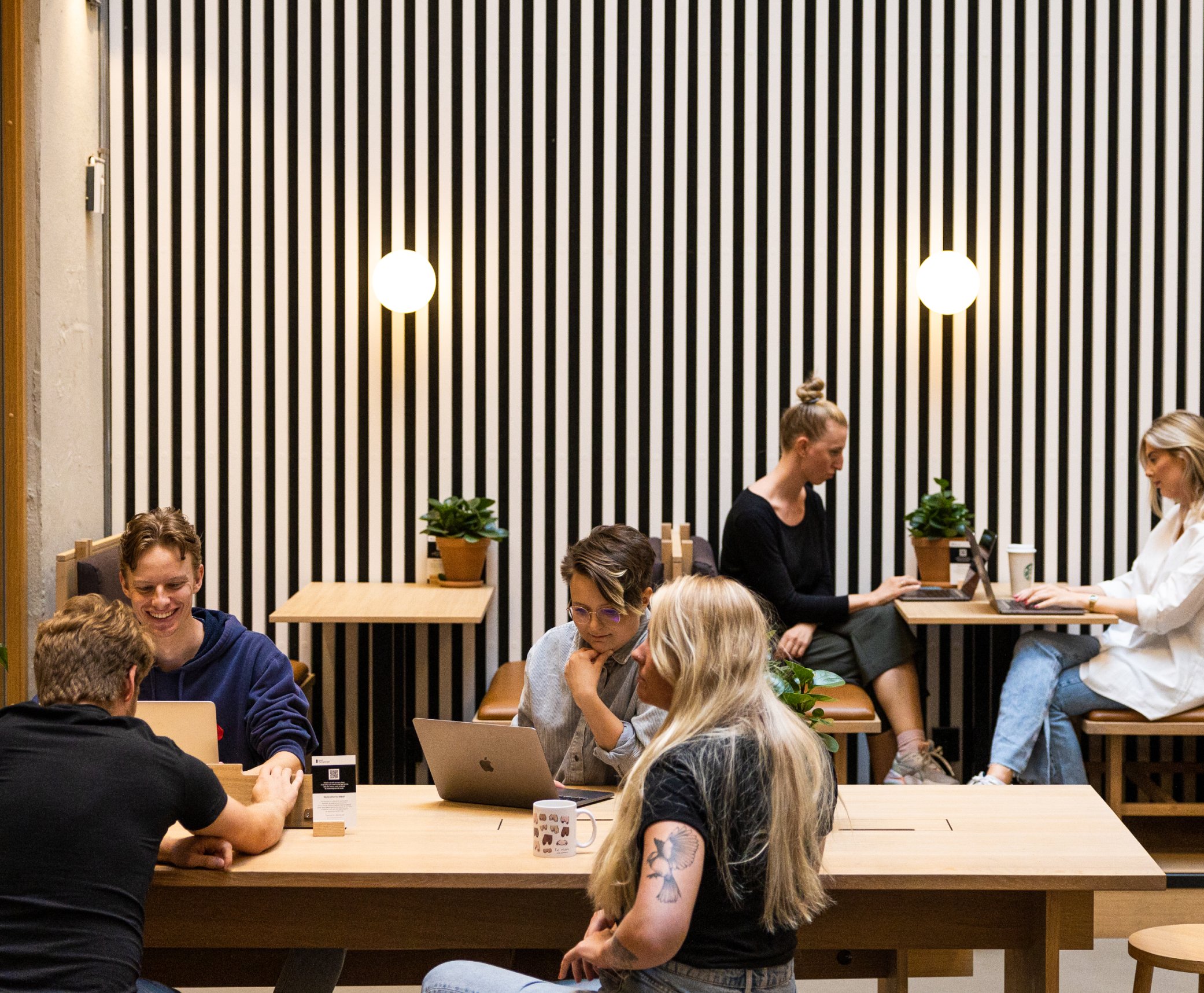 en gruppe mennesker sitter ved bord med bærbare datamaskiner foran en stripete vegg.