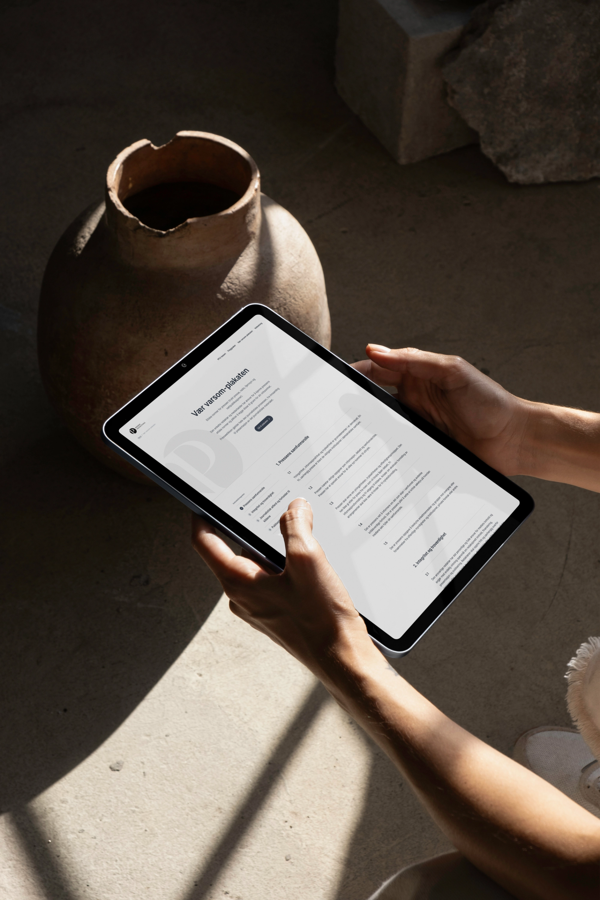 Hands holding a tablet displaying a document, with a rustic pot and rocks in the background.