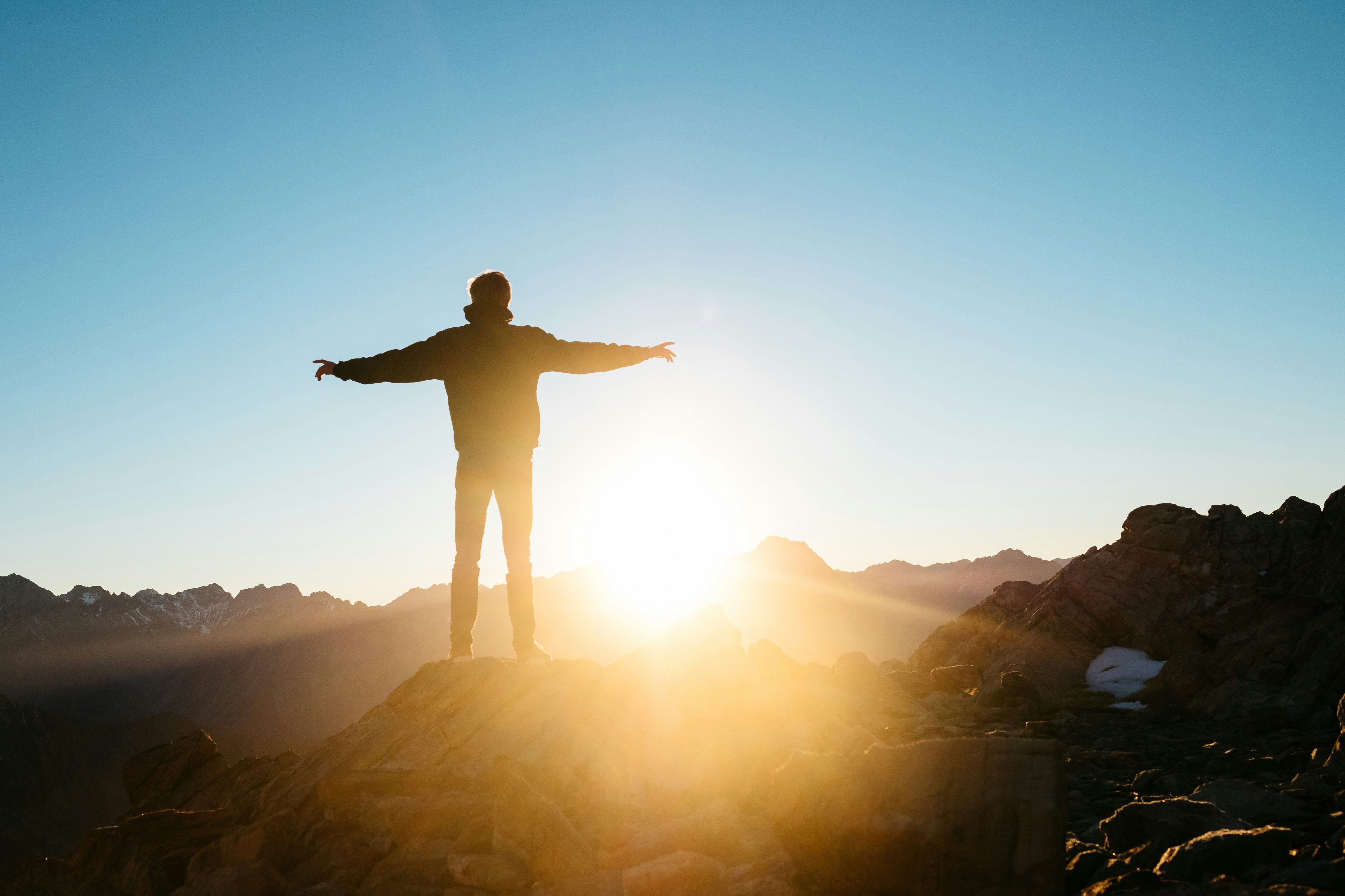 a man is standing on top of a mountain with his arms outstretched at sunset .