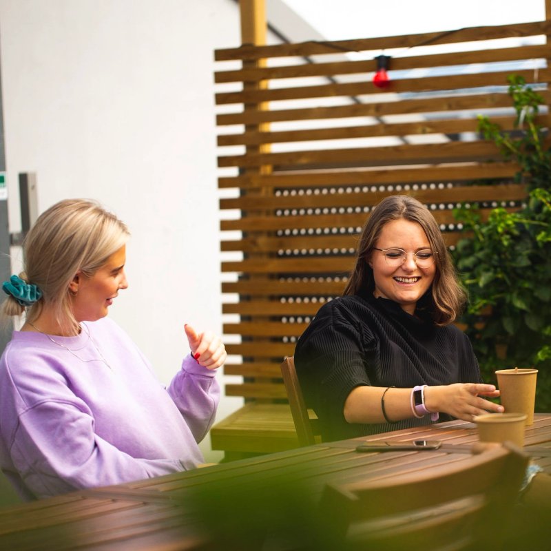 Caroline og Vilde i Kult Byrå sitter på et bord, drikker kaffe og prater