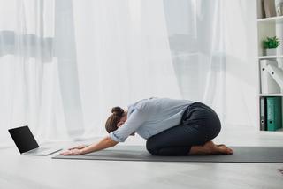 Dame gjør yoga på gulvet på kontoret, med laptop foran seg. Foto