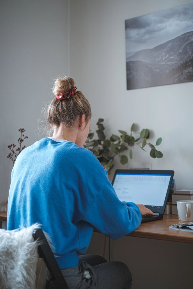 Jente sitter på hjemmekontor iført collegegenser og jobber på en laptop. Foto