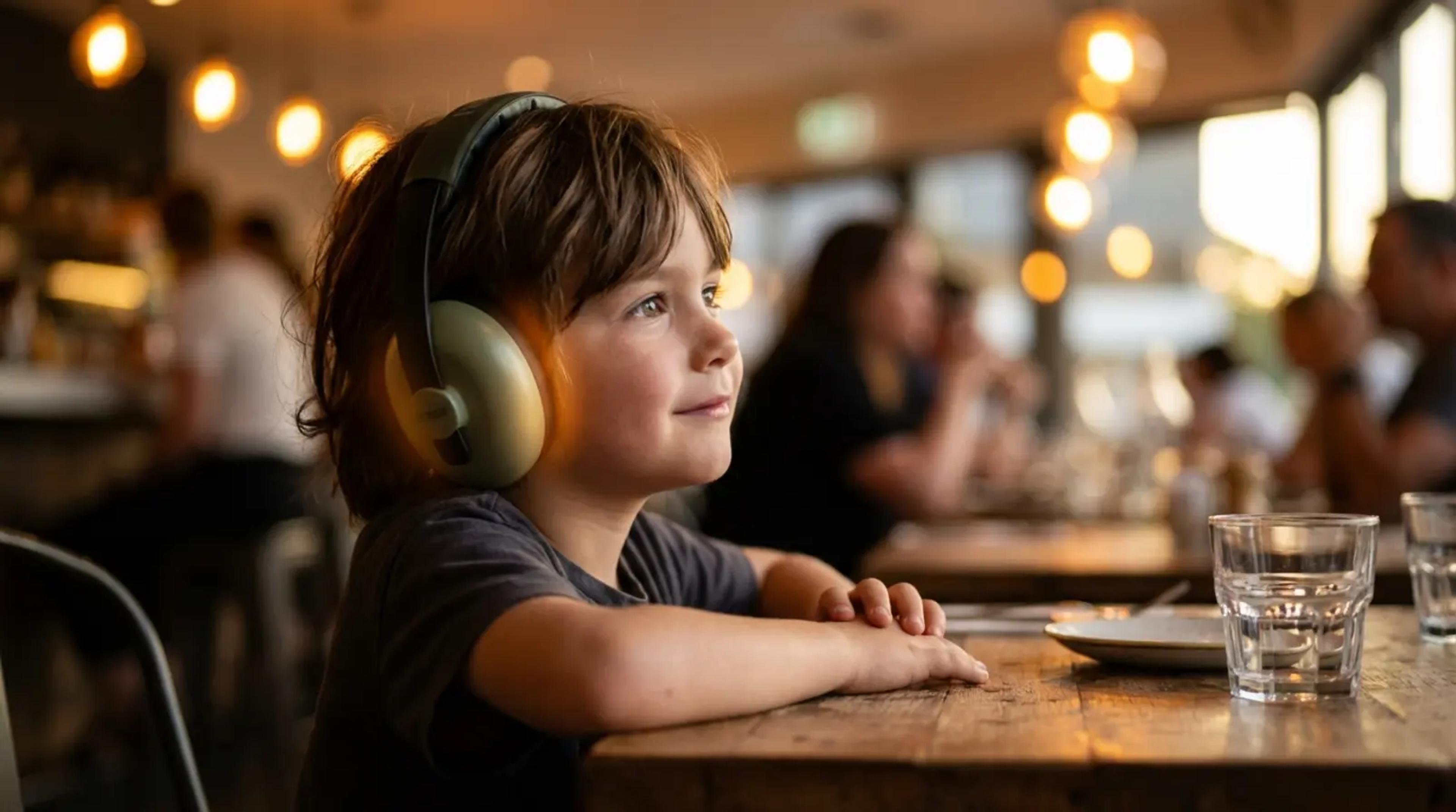 A young child sitting calmly at a busy restaurant table wearing headphones, looking engaged and imaginative while listening to an audio story instead of using a screen.