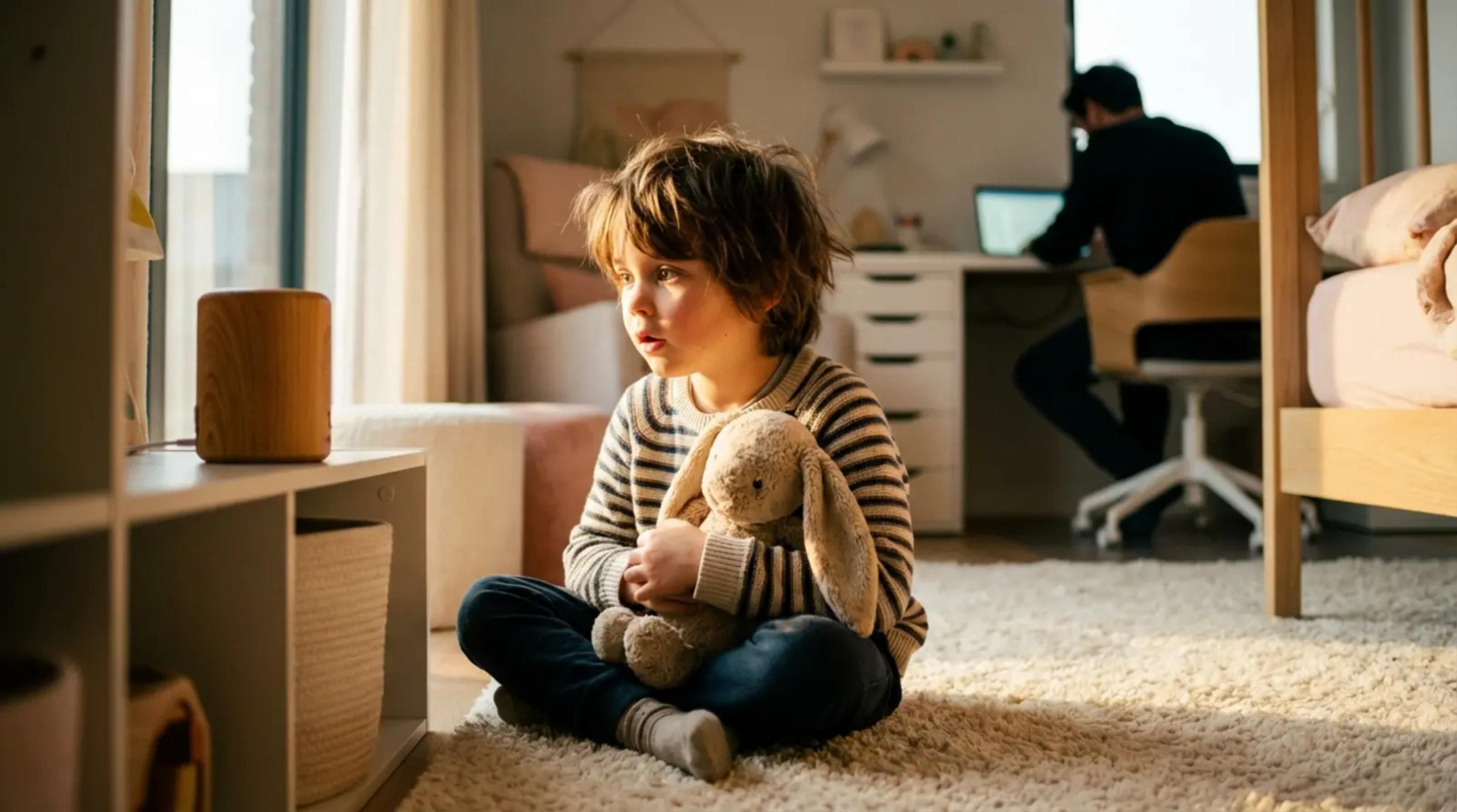 A young child sitting on a rug playing with a stuffed toy while a parent works in the background