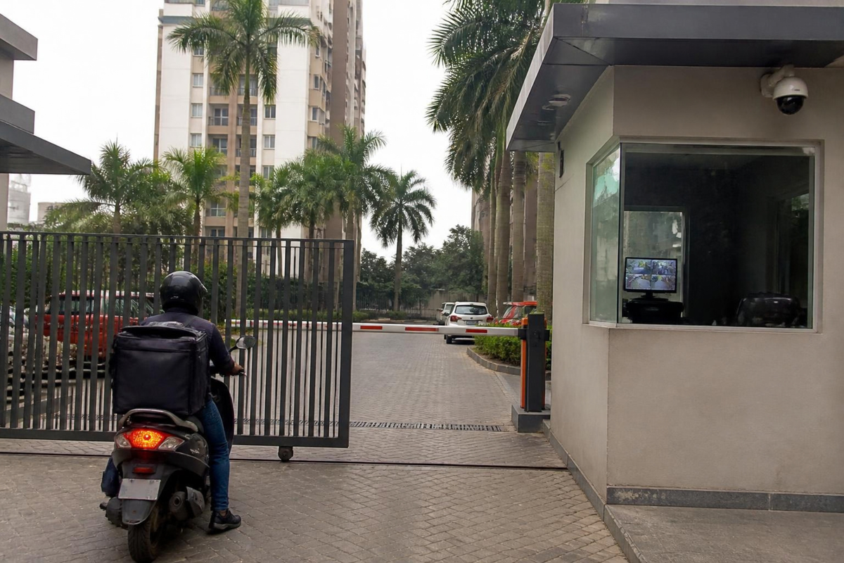 Delivery rider at a housing society gate with a boom barrier and guard cabin.