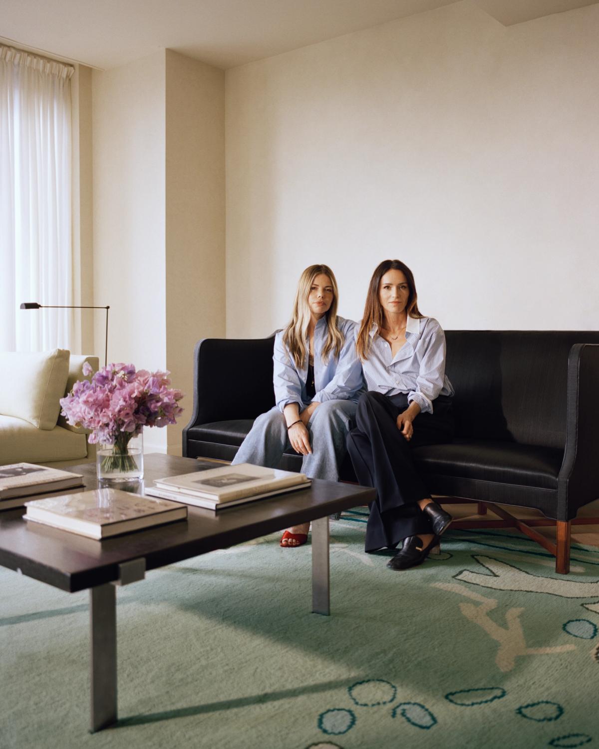 Portrait of creative director Alex Eagle and interior designer Sophie Hodges, seated on a black sofa in a modern living room. The space is styled with a dark wood coffee table stacked with books and the Giverny – Green rug from their collection with Nordic Knots.