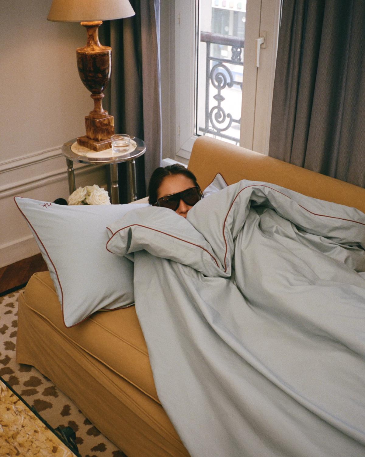 Diana Bartlett entspannt auf einem beigen Sofa, eingehüllt in die Nordic Knots Percale Duvet Cover in Shirt Blue/Red, in einem gemütlichen und eleganten Interieur mit Beistelltisch, Lampe und sanftem Tageslicht.