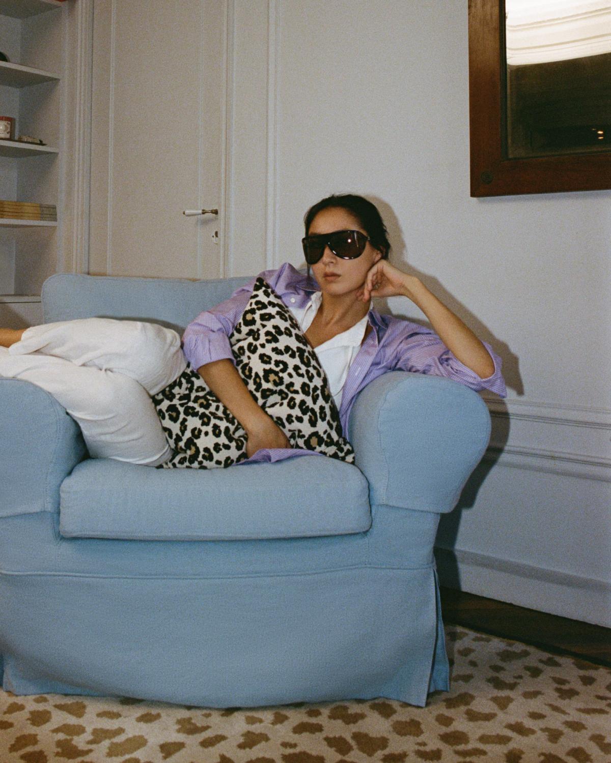 Diana Bartlett sitzt in einem hellblauen Sessel und hält das Leopard-Kissen in Beige, mit dem Gild 03 – Leopard-Teppich darunter, in einem sanft beleuchteten Wohnzimmer.