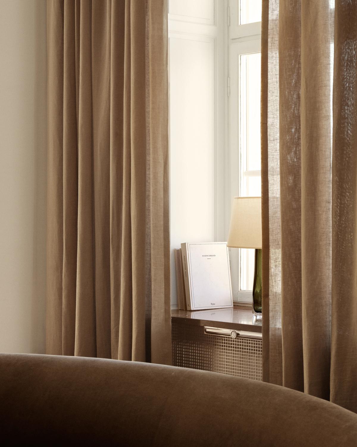 Soft and airy interior detail with The Sheer Linen – Hazelnut curtain framing a window, styled with a table lamp and books on the windowsill.