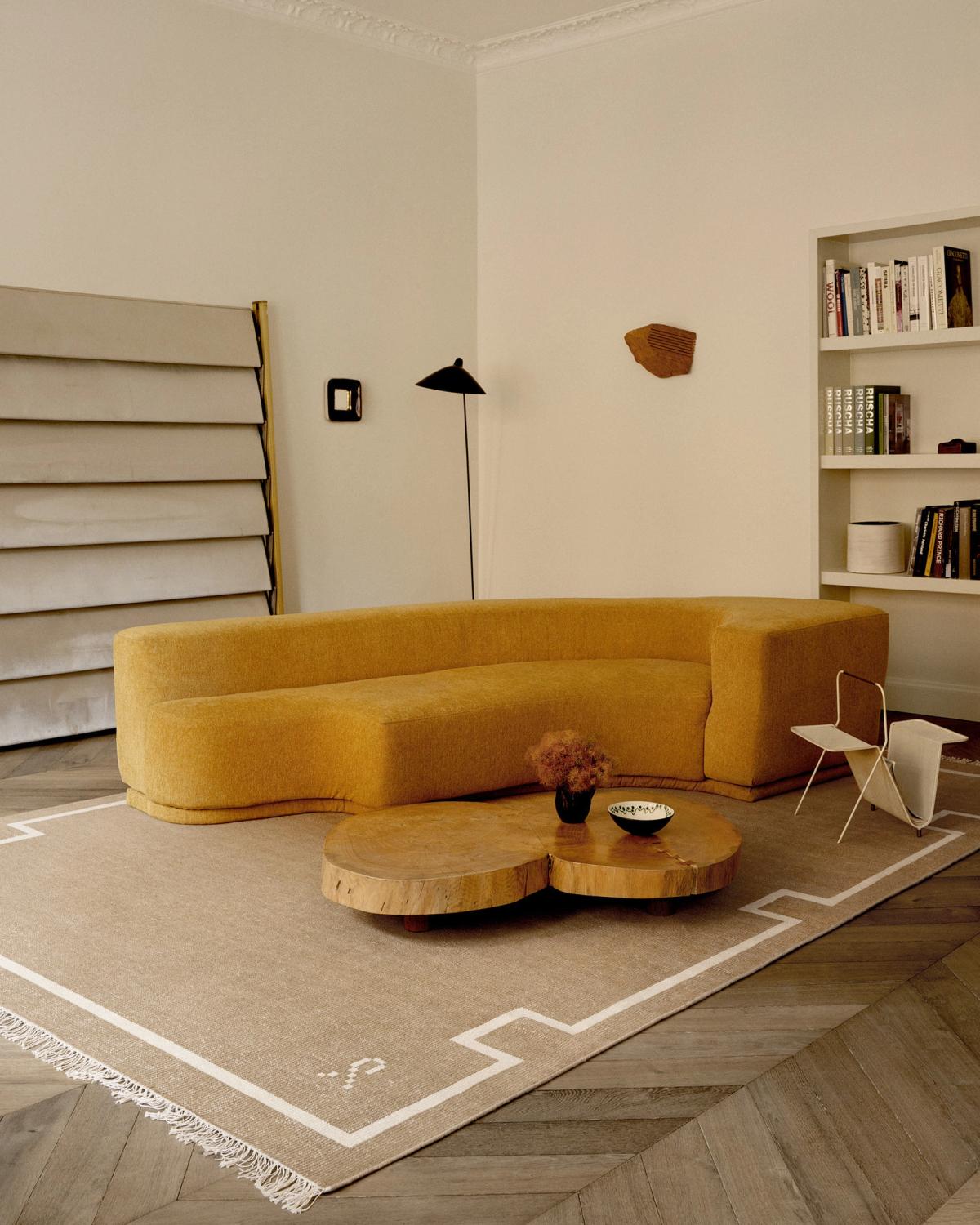 Der Elder 05-Teppich in Cream wird in einem modernen Wohnzimmer mit einem geschwungenen senffarbenen Sofa, Holztischen und einer geometrischen Stehlampe gezeigt und schafft eine warme und minimalistische Atmosphäre.