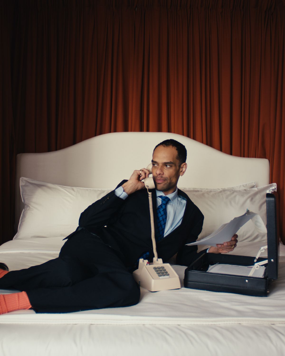 Raven Smith, auf einem Bett mit frischer weißer Bettwäsche in der 130 Mount Street in London liegend, eingerahmt von Burnt Red Vorhängen, mit einem Telefon und geöffnetem Aktenkoffer neben sich.