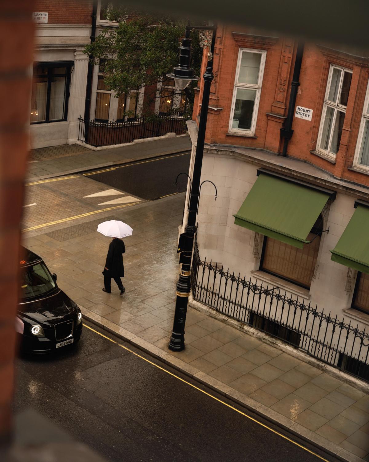Ein Blick aus dem Nordic Knots-Geschäft in der Mount Street in London zeigt eine regnerische Straßenszene mit einer Person, die unter einem weißen Regenschirm an roten Backsteingebäuden und grünen Markisen vorbeigeht.