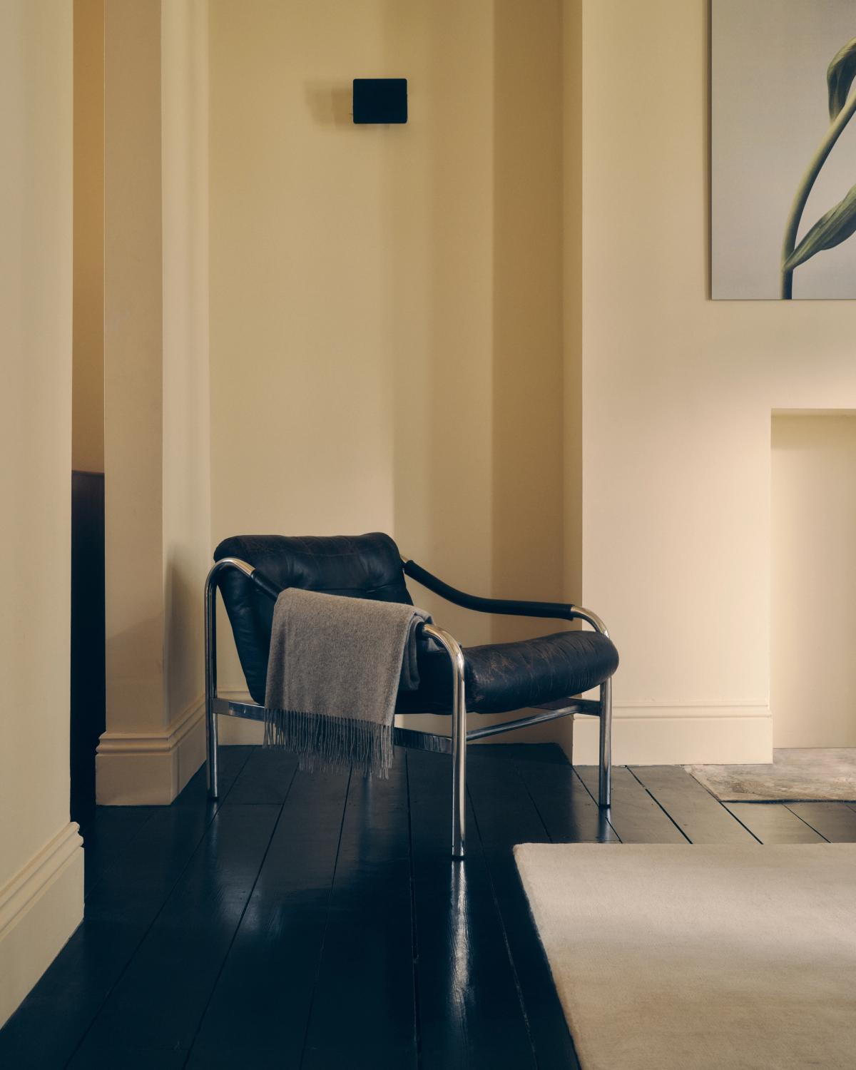 Flauschiger Wollteppich Grand in Dusty White, abgebildet auf einem schwarzen Holzboden mit einem schwarzen Ledersessel daneben.