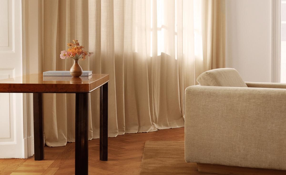 Die Sheer-Vorhänge in Pale Sand sind in einem sonnenbeschienenen Wohnzimmer mit einem beigen Sofa, einem Holztisch und Parkettboden zu sehen und schaffen eine weiche, natürliche Atmosphäre.