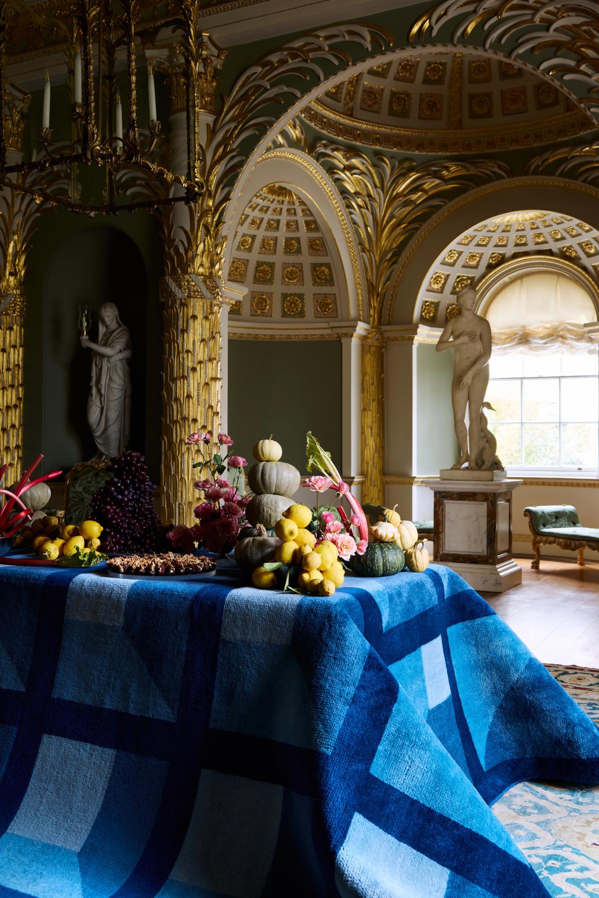Plush wool rug Garden Maze in Blue pictured draped over a table with a decadent spread of fruits and flowers in a museum like setting.
