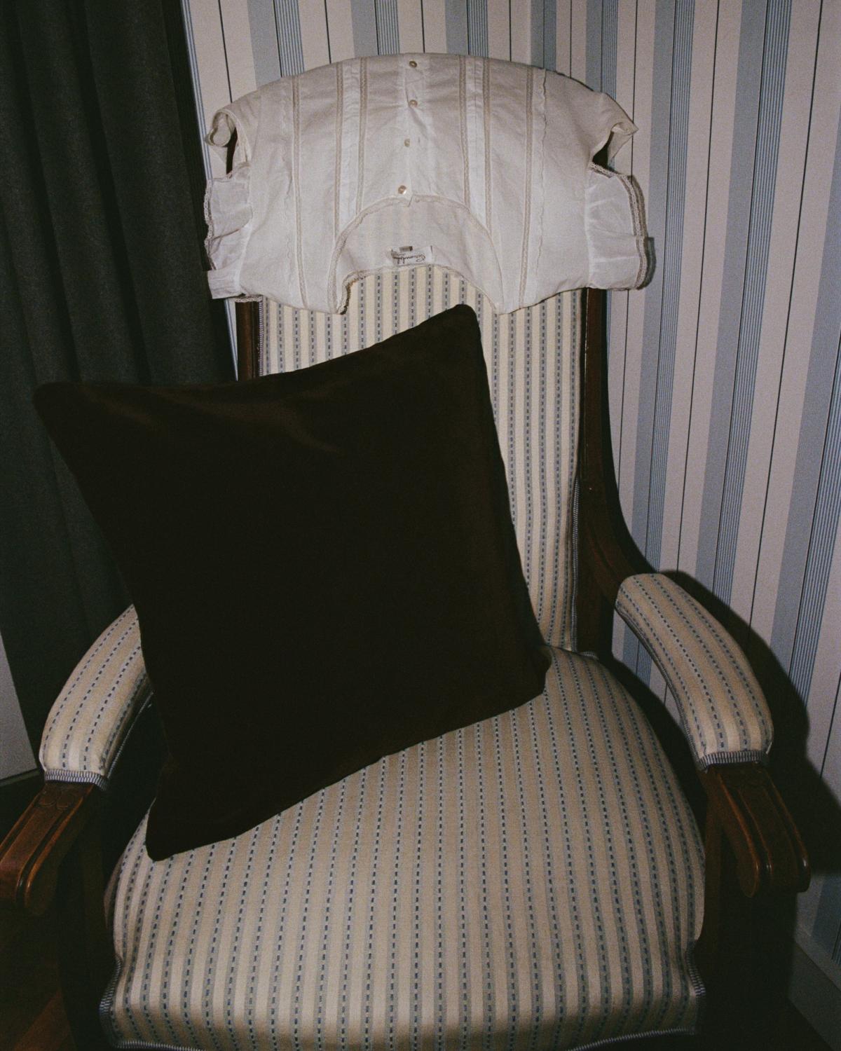 Das Teddy Velvet-Kissen in Walnut liegt auf einem gestreiften, gepolsterten Sessel, über dessen Rückenlehne ein weißes Hemd hängt, vor einer gestreiften Tapete.