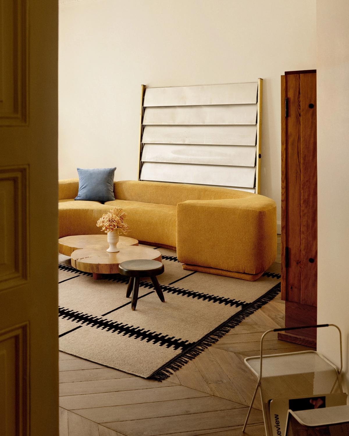 The Elder 06 rug in Black is shown in a contemporary living room with a curved mustard sofa, wooden coffee tables, and a soft blue cushion, creating a warm and modern atmosphere.