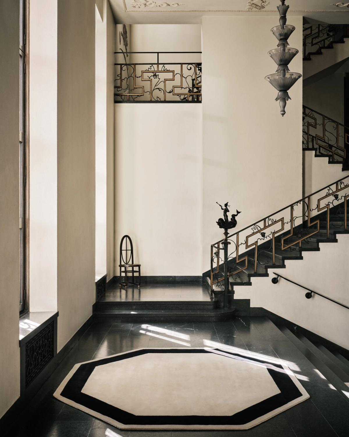 Grace 05 – Octagon — ein achteckiger Teppich in neutralem Beige, eingefasst von einer markanten schwarzen Bordüre, fotografiert im Stockholmer Konzerthaus mit skulpturalen Treppengeländern und sanftem natürlichen Licht.