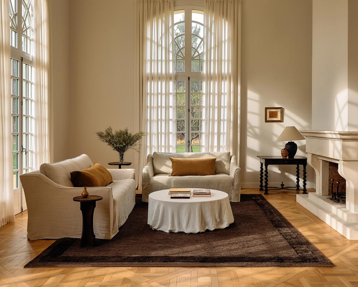 Vue large du tapis Jute Frame en coloris Dark Brown dans un salon lumineux, ancrant l’espace avec une cheminée, du mobilier moderne et de grandes fenêtres pour une ambiance aérée.