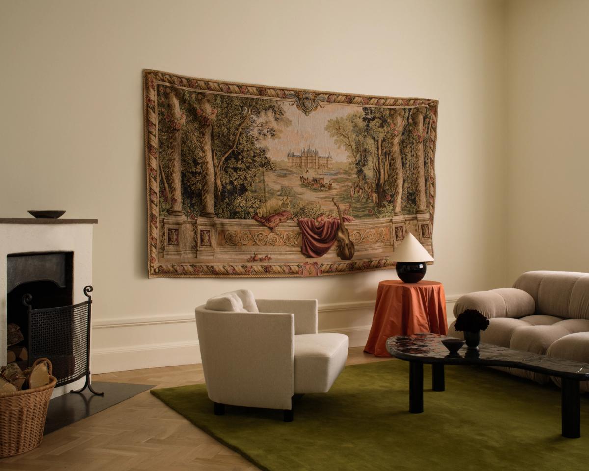 The plush wool rug Grand in Milano Green showcased in a beautiful living room, paired with a white armchair and a beige sculptural sofa. The rug also sits underneath a black asymmetrical coffee table.