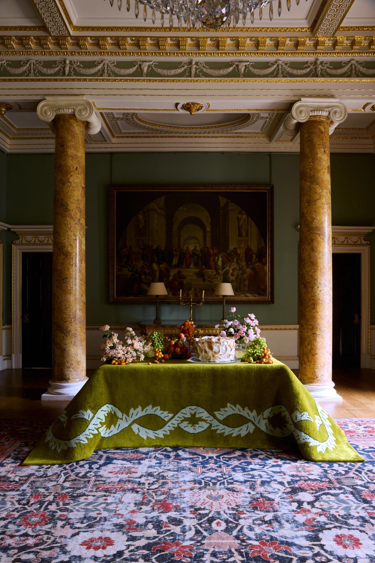 Plush wool rug Climbing Vine in Olive pictured draped over a table with a decadent cake and flowers in a museum like room.